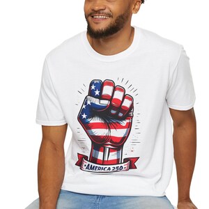 May include: White t-shirt featuring a graphic of a clenched fist with the American flag design. The fist is red, white, and blue, with stars and stripes. A banner below the fist reads "AMERICA 250".