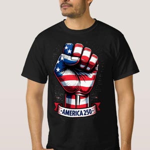 May include: Black t-shirt featuring a graphic of a clenched fist painted with the American flag design. The fist is red, white, and blue, with stars on the blue section. A banner below the fist reads "AMERICA 250".