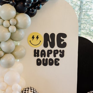 May include: A white backdrop with black and white checkered balloons, white and gray balloons, and a yellow smiley face with lightning bolts. The backdrop has black letters that say "ONE HAPPY DUDE".