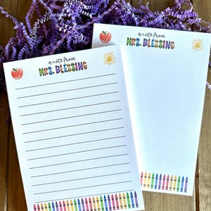 May include: Two white notepads with a colorful border of crayons at the bottom. The top of each notepad has a colorful title that says "a note from MRS. BLESSING" with a red apple and a yellow sun on each notepad.
