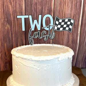 May include: A white frosted cake with a decorative topper that reads "TWO fast" with a checkered flag. The cake is on a white cake board, set against a wooden background.