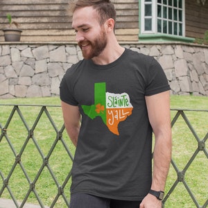 Könnte beinhalten: Dunkelgraues T-Shirt mit einem grünen und orangefarbenen Texas-Umriss, einem Kleeblatt und den Worten "Sláinte Y'all" in Weiß und Grün. Das T-Shirt hat einen Rundhalsausschnitt und kurze Ärmel.