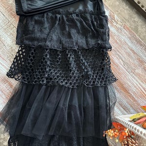 May include: A black skirt featuring multiple horizontal layers of fabric. The design includes a top tier of solid black fabric, followed by a layer of black lace, a layer of black fishnet mesh, and a bottom layer of black tulle over a lace hem.