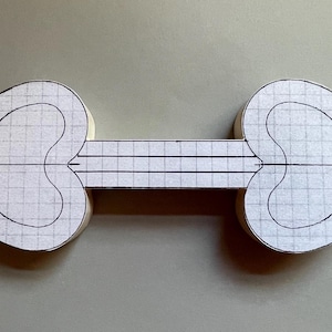 Pode incluir: Um design de artesanato em papel com dois elementos em forma de coração conectados por uma ponte retangular. Os corações e a ponte são brancos com um padrão de grade e contorno preto, em um fundo neutro.