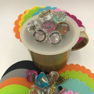 May include: A collection of colorful glass charms with inspirational words like "Hope", "Dream", "Happiness", "Peace", and "Strong" are displayed in a white mug. The charms are surrounded by colorful silicone coasters.