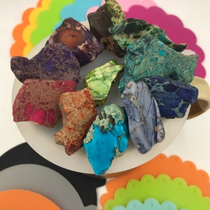 May include: A collection of colorful, irregularly shaped, polished stones with a marbled pattern. The stones are arranged on a white surface with a rainbow of silicone coasters in the background.