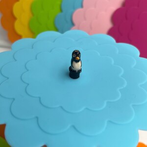May include: A set of colorful silicone lids with a penguin-shaped knob. The lids are in various colors, including orange, yellow, green, pink, and purple. The lids are designed to fit on cups and bowls.