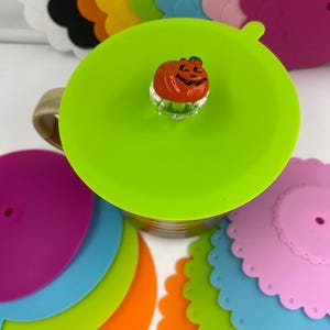 May include: A collection of colorful silicone cup lids in various shades, including green, orange, blue, pink, and white. A green lid with a pumpkin-shaped topper sits atop a mug. The lids have a scalloped edge design.