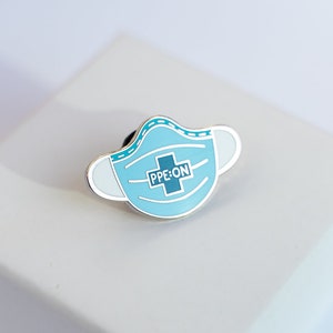 May include: A blue enamel pin shaped like a face mask with the text "PPE ON" and a white cross inside. The pin is on a white square box.