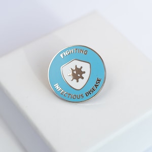Op de afbeelding: Een blauwe en witte emaille pin met een schildontwerp. Het schild bevat een gestileerd beeld van een virus en de tekst "Fighting Infectious Disease".