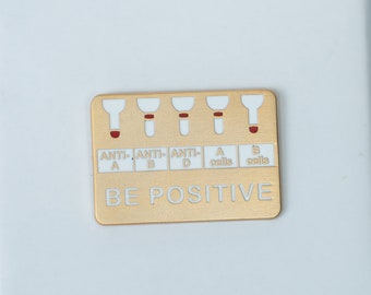 May include: A gold-colored metal pin with a white background and the text "BE POSITIVE". The pin features a stylized image of a blood test result with five lines, each with a red dot at the end.