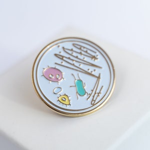 May include: A gold-coloured pin with a white background depicting a petri dish with a pink, yellow, and turquoise bacteria and a few lines of what appears to be a scientific drawing.