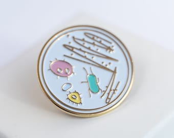 May include: A gold-coloured pin with a white background depicting a petri dish with a pink, yellow, and turquoise bacteria and a few lines of what appears to be a scientific drawing.