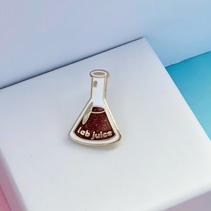 Könnte beinhalten: Eine goldfarbene Emaille-Anstecknadel in Form eines Bechers mit einer roten Flüssigkeit darin. Der Becher trägt den Text "lab juice".