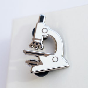 May include: A white metal microscope pin with black details. The pin is on a white background.