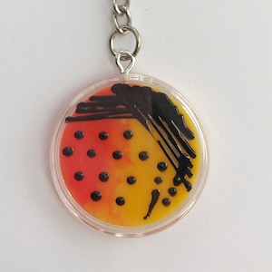 Puede incluir: Un llavero con un dije redondo de plástico transparente con fondo rojo y amarillo y un diseño abstracto negro. El dije está unido a una cadena plateada y un anillo plateado.