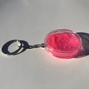 Puede incluir: Llavero de placa de Petri rosa con tapa transparente. La placa contiene un medio de agar rosa con rayas y puntos blancos. El llavero tiene un anillo y una cadena plateados. El texto '@shopmicrobi' es visible en la placa.