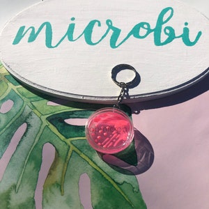 Puede incluir: Llavero de slime rosa y blanco con una carcasa de pl&aacute;stico transparente. El llavero est&aacute; unido a un llavero plateado. El llavero cuelga de un cartel ovalado blanco con la palabra "microbi" escrita en turquesa.