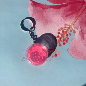 Puede incluir: Llavero con una placa de Petri con bacterias rosas y un anillo de llave plateado. El llavero est&aacute; sobre un fondo azul con una flor rosa de acuarela.