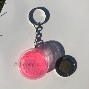 Puede incluir: Un llavero de placa de Petri rosa con una cadena y un anillo plateados. La placa de Petri tiene un agar rosa con rayas blancas y puntos rojos. El texto "@shopmicrobi" es visible en la placa de Petri.