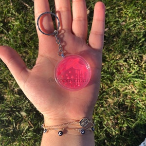Puede incluir: Un llavero de placa de Petri rosa con bacterias rojas y blancas en su interior. El llavero est&aacute; unido a una cadena y un anillo plateados. El llavero se sostiene en una mano con una pulsera dorada con dijes de ojo azul.