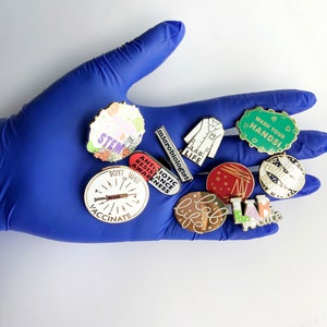 May include: A blue gloved hand holds a collection of enamel pins with various science and medical themes. The pins feature phrases like "Lab Life", "Don't Wait Vaccinate", "Wash Your Hands", and "Antibiotic Resistance Awareness".