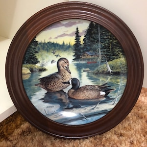 May include: A framed ceramic plate with a painting of two blue-winged teal ducks in a forest setting. The plate has a brown wooden frame and the artist's signature "Jerner" is visible in the bottom right corner.