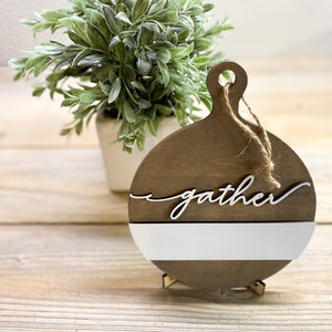 May include: A wooden round sign with a white stripe and the word "gather" in white script. The sign is on a small wooden stand and has a twine loop for hanging.