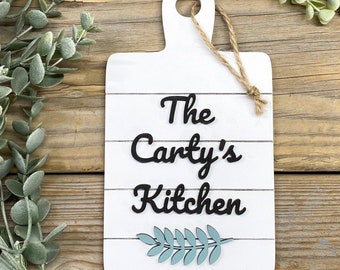 Könnte beinhalten: Ein weißes, hölzernes Küchenschild mit dem Text "The Carty's Kitchen" in schwarzen Buchstaben. Unter dem Text befindet sich ein blaues, gemaltes Blattdesign.