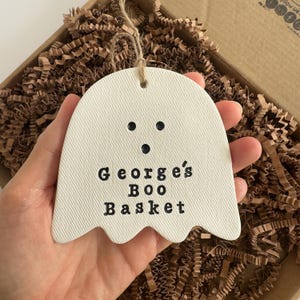 May include: A cream-coloured ghost-shaped ornament with black dot eyes and mouth. The text "George's Boo Basket" is printed in black. The ornament is suspended by a natural twine string and is presented in a cardboard box with brown paper filler.