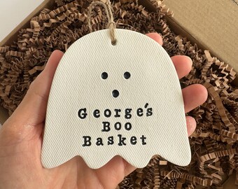 May include: A cream-colored ghost-shaped ornament with black dot eyes and mouth. The text "George's Boo Basket" is printed in black. The ornament is suspended by a natural twine string and is presented in a cardboard box with brown paper filler.