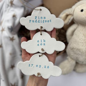 May include: Three white ceramic cloud-shaped ornaments with blue text. The top cloud reads "Finn Puddifoot", the middle cloud reads "6 lb 4 oz", and the bottom cloud reads "17.03.26". Each cloud is strung with twine. A stuffed lamb is in the background.