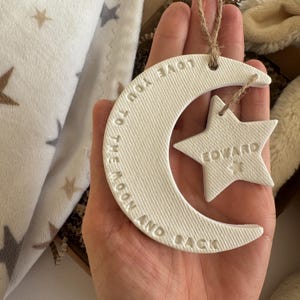 May include: A white ceramic moon and star ornament. The crescent moon has the words "LOVE YOU TO THE MOON AND BACK" embossed on it. A star-shaped ornament is attached, with the name "EDWARD" embossed. The ornament is held by a string.