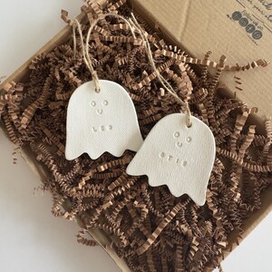 May include: Two white, ghost-shaped ornaments made of a textured material, each featuring a simple stamped face and the names Leo and Otis. The ornaments are suspended by thin twine strings and are nestled within a brown cardboard gift box.