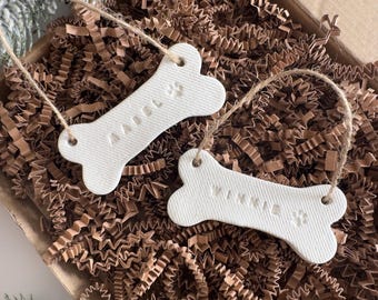 May include: Two white bone-shaped ornaments, each with a name embossed: "MABEL" and "WINNIE". A paw print is also visible on each. They hang by twine loops, presented on brown crinkle paper within a cardboard box.