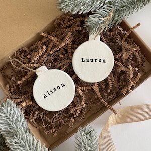 May include: Two cream-coloured, round Christmas baubles with the names "Alison" and "Lauren" printed in black. The baubles are in a cardboard box filled with brown shredded paper, with faux evergreen branches and a gold ribbon.