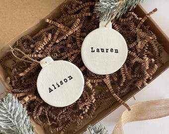 May include: Two off-white, round Christmas ornaments with the names "Alison" and "Lauren" printed in black. The ornaments are in a cardboard box filled with brown crinkle paper, with faux evergreen branches and a gold ribbon.