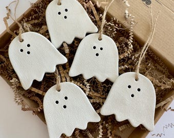 May include: Five white, ghost-shaped ornaments made of a textured material, each featuring three small black holes for eyes and a mouth. Each ghost is suspended by a thin, natural-colored jute twine loop, arranged inside a brown cardboard box.
