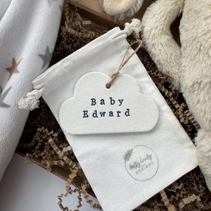 May include: A white cloud-shaped tag with the text "Baby Edward" in navy blue, attached to a white drawstring bag. The bag is inside a brown box with shredded paper. A soft, cream-coloured plush toy is visible in the background.