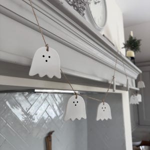 May include: A garland of white ghost decorations with black dot eyes, strung on twine. The ghosts are hanging in front of a painted grey mantelpiece, ideal for Halloween. The mantelpiece is above a tiled surface.