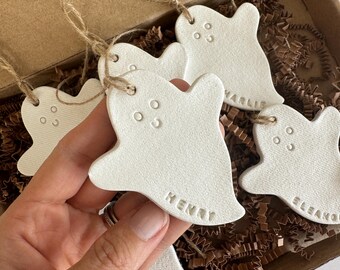 May include: A collection of white ghost-shaped ornaments with jute twine hangers. Each ornament has three small circular eye indentations and a name. The ornaments are in a cardboard box with brown paper filler.