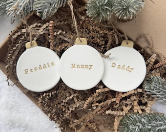 May include: Three circular white ornaments with gold-colored caps and twine loops. Each ornament features a name embossed in gold lettering: Freddie, Mummy, and Daddy. The ornaments are arranged in a brown box filled with brown paper filler.