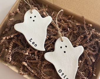 May include: Two white ghost-shaped ornaments with black dot eyes and the names "Leo" and "Otis" printed on them. The ornaments are hanging by twine strings and are in a cardboard box filled with brown paper filler.