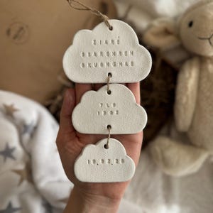 May include: Three white cloud-shaped ornaments suspended by twine. Each cloud features embossed text. The top cloud has three lines of text. The middle cloud has weight measurements of 3.2 kg. The bottom cloud has a date.