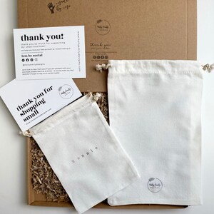 May include: A brown cardboard shipping box containing two off-white cotton drawstring pouches, one with the name Ronnie printed on it and the other with a logo. Two white thank you cards with black text are included, one reading thank you!