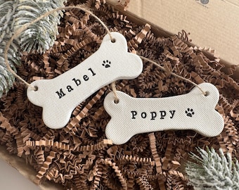 May include: Two off-white bone-shaped ornaments with a textured surface, each featuring a jute string for hanging. One ornament is stamped with the name Mabel and a paw print, while the other is stamped with the name Poppy and a paw print.
