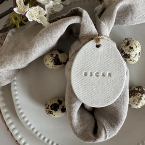 May include: A light grey linen napkin shaped like a bunny, with a white ceramic name tag that reads "OSCAR". The tag is tied with twine. Speckled quail eggs and a white plate complete the Easter table setting.