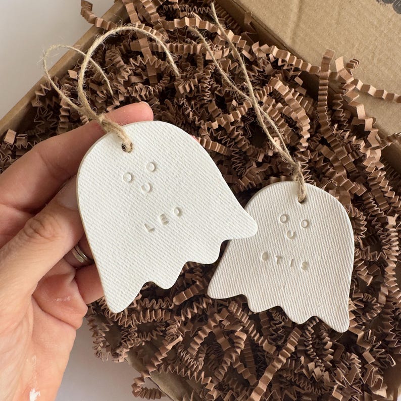 May include: Two white, ghost-shaped ornaments made of a textured material, each featuring a simple stamped face and a name. One ghost is stamped with the name LEO and the other with the name OTIS. Both ornaments hang from thin, natural jute twine.