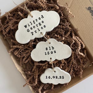 May include: Three cloud-shaped ceramic ornaments with jute twine, in a cardboard box with brown paper filler. The top ornament reads "Wilfred George Price." The middle ornament shows "8 lb 15 oz." The bottom ornament displays "16.02.25."