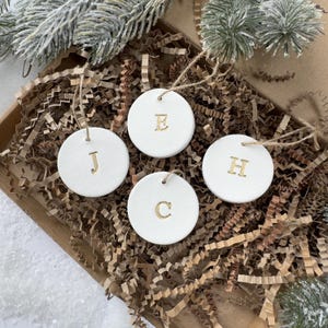 May include: Four white, circular Christmas decorations, each with a gold letter: J, E, C, and H. They hang from twine and are presented in a brown cardboard box with shredded paper. Evergreen branches are visible in the background.